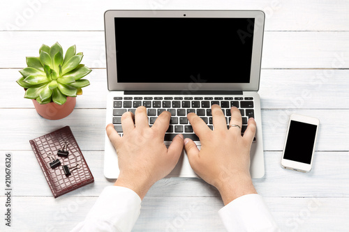Conceptual workspace or business concept. Laptop computer with male hands, plant in a pot, notebook with paper clips and modern mobile phone on white wooden background.
