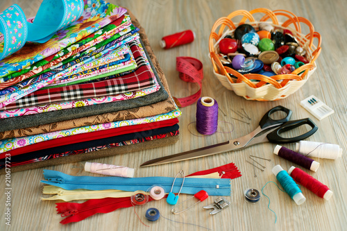 Items for sewing clothes on the table. A stack of fabric, a centimeter tape, tailor's scissors, a zipper lock, a basket with small objects for sewing clothes.