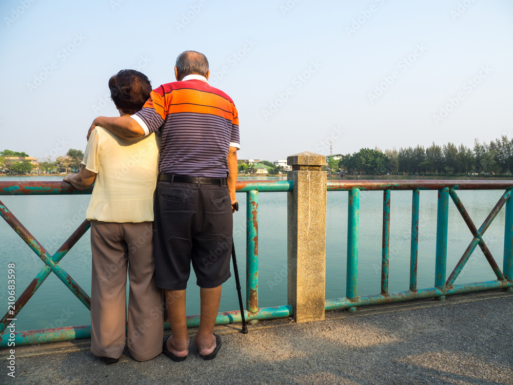 Happy romantic senior asian couple stand on the bridge in front of the lake. Husband stands with his wife. Concept of senior couple and take care of each other
