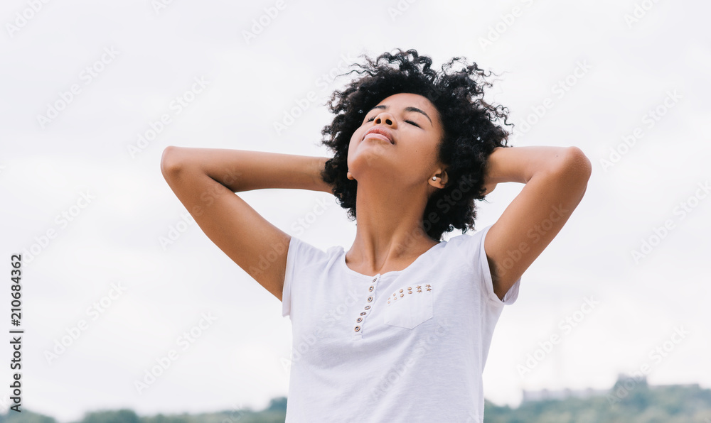 Image of happy african american woman having rest outdoors in the park, with hands on raised head and closed eyes, feel free. Beautiful young dark-skinned female enjoy nature and healthy life. People