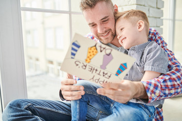 Portrait of happy young father hugging cute son and celebrating Fathers day together sitting by window at home, copy space