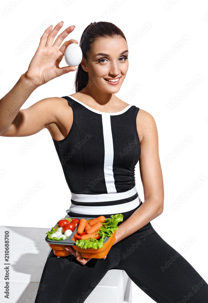 Smiling sporty woman holding container with healthy nutrition and showing chicken egg. Photo of attractive latin woman in fashionable sportswear on white background. Healthy lifestyle