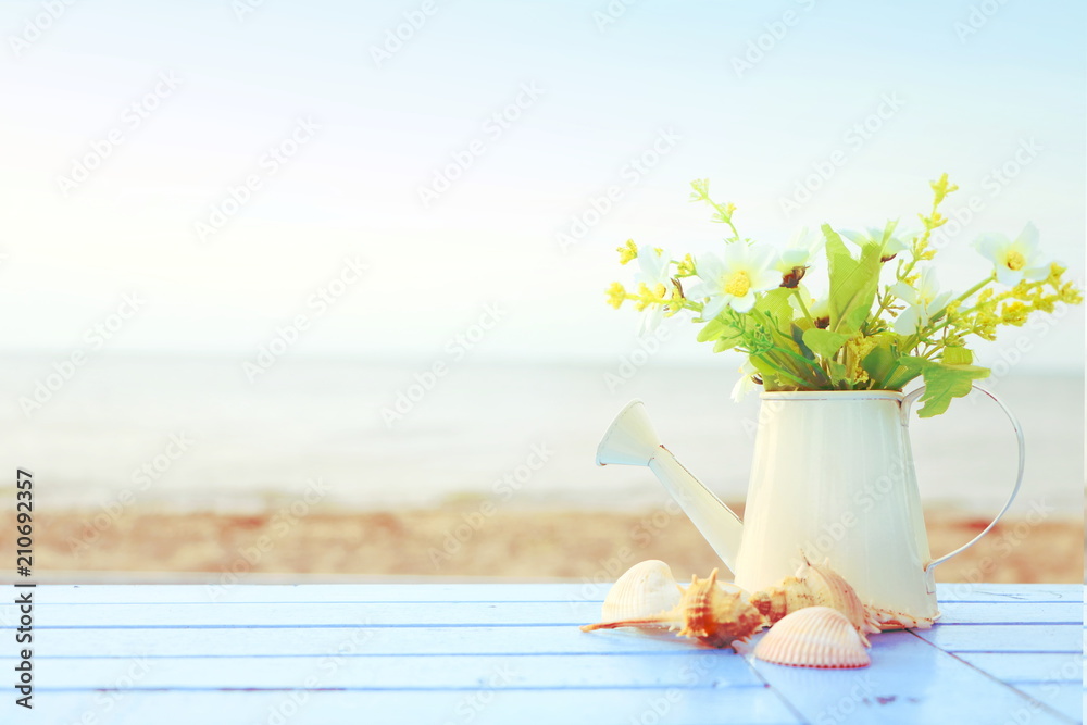 many little white flower in white watering vase with brown shells on blue wooden table on blur sea view background, concept summer on the beach, feeling soft and relax, vintage tone