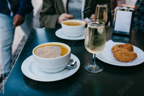 A person is going to eat traditional Portuguese food - soup and patty with a cod called Pastel and drink a glass of white wine in a street cafe