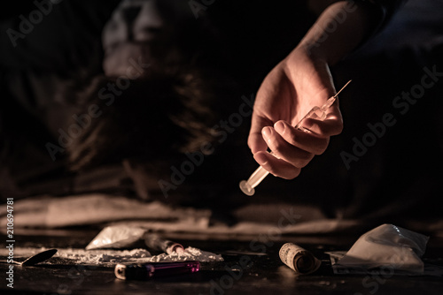 Male junkie hand holding drug injection syringe while lying near heroin powder, spoon and cigarette lighter for heroin cooking and money on dark floor. Hard drug addiction concept