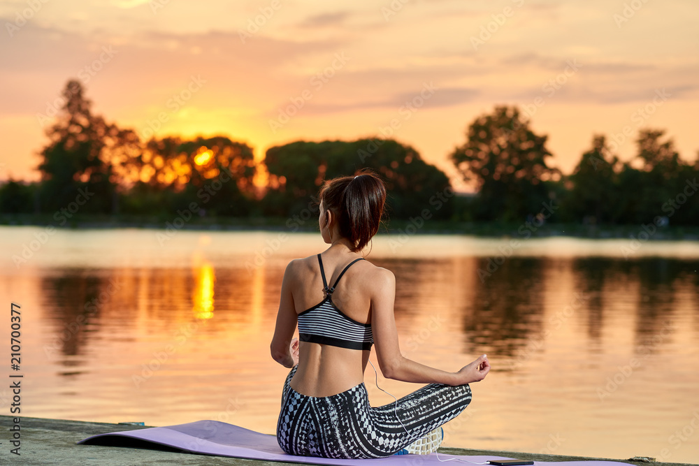 Backview of young sportswoman sitting in lotus position near city lake in evening on sunset. Stylish sporswear. Maintaining healthy lifestyle. Sport, yoga, fitness. Girl having fit, beatiful body.
