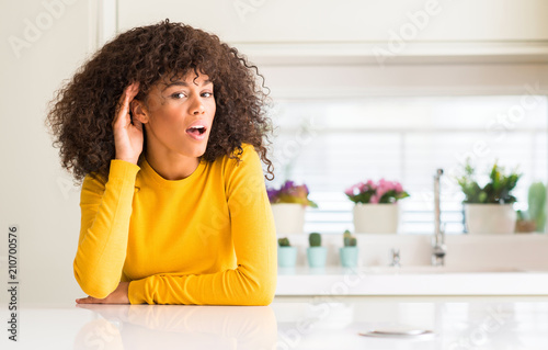 African american woman wearing yellow sweater at kitchen smiling with hand over ear listening an hearing to rumor or gossip. Deafness concept.