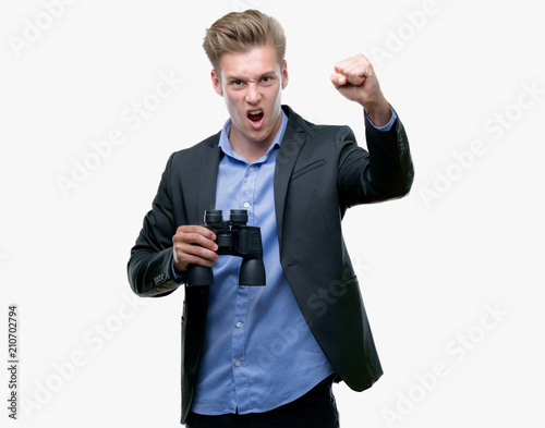 Young handsome blond man looking through binoculars annoyed and frustrated shouting with anger, crazy and yelling with raised hand, anger concept