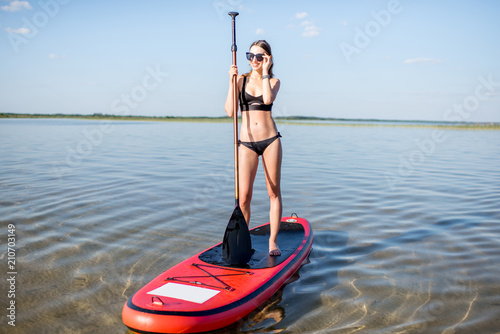 Portrait of a young woman in black swimsuit paddleboarding on the lake with calm water during the sunset © rh2010