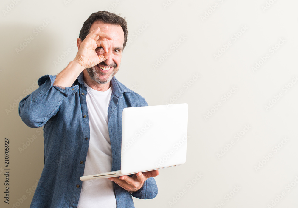Senior man using laptop computer with happy face smiling doing ok sign with hand on eye looking through fingers Senior man using laptop computer with happy face smiling doing ok sign with hand on eye looking through fingers