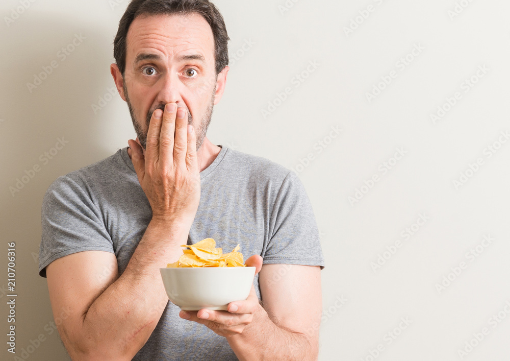 Senior man eating potato chips cover mouth with hand shocked with shame for mistake, expression of fear, scared in silence, secret concept Senior man eating potato chips cover mouth with hand shocked with shame for mistake, expression of fear, scared in silence, secret concept