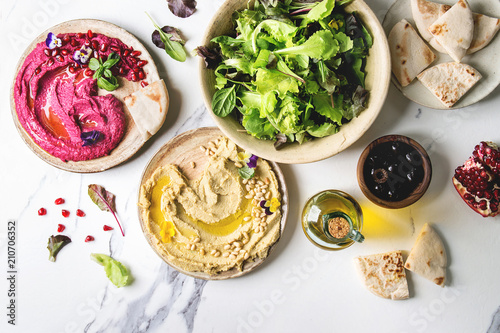 Variety of homemade traditional and beetroot spread hummus with pine nuts, olive oil, pomegranate served on ceramic plates with pita bread and green salad on white marble background. Flat lay, space.