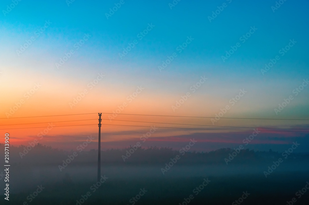 Evening thick fog over field and forest. Landscape of the evening sky with orange and blue sky and fog. Telegraph pole in the fog.