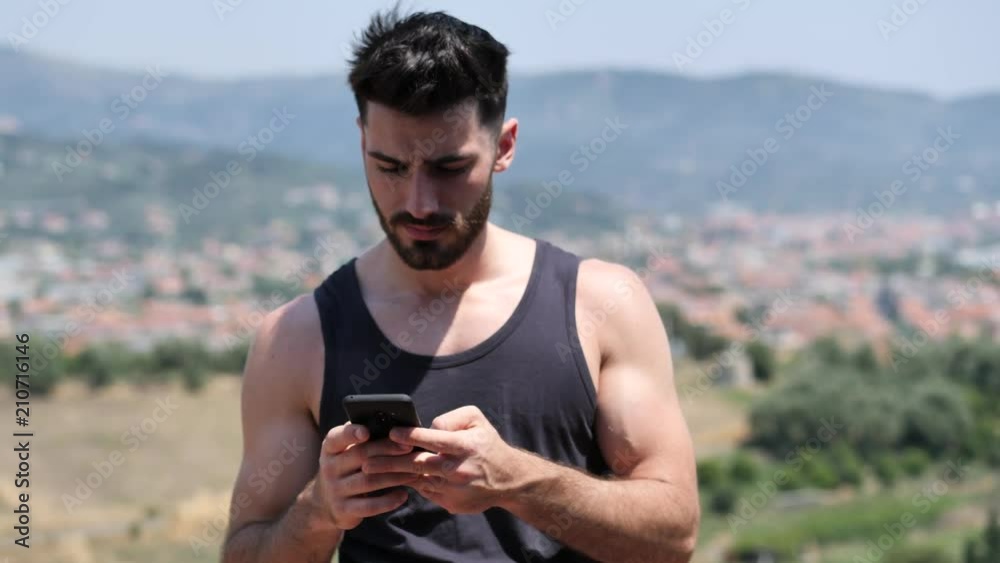 Young handsome man using smartphone, typing text message to someone while standing on a balcony at the seaside over countryside landscape Young handsome man using smartphone, typing text message to someone while standing on a balcony at the seaside over countryside landscape