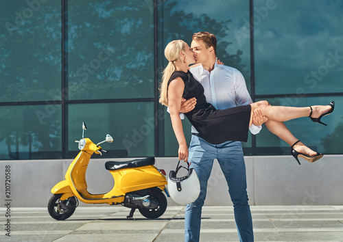 Happy attractive couple in love - handsome stylish guy holds on hands and kiss his charming blonde girlfriend near yellow classic Italian scooter against a skyscraper. Together in love, date, romance