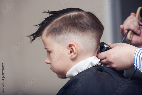 Barber is shaving a hair to Caucasian boy in barbershop.
