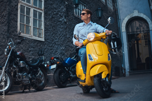 Young stylish guy dressed in a in a white shirt and jeans ride on yellow classic italian scooter on an old Europe street in the evening. © Fxquadro