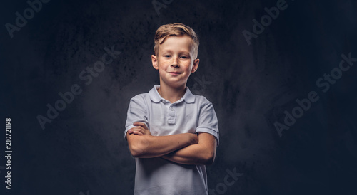 Cute little boy dressed in a white t-shirt standing with crossed arms in a studio. Isolated on dark textured background.
