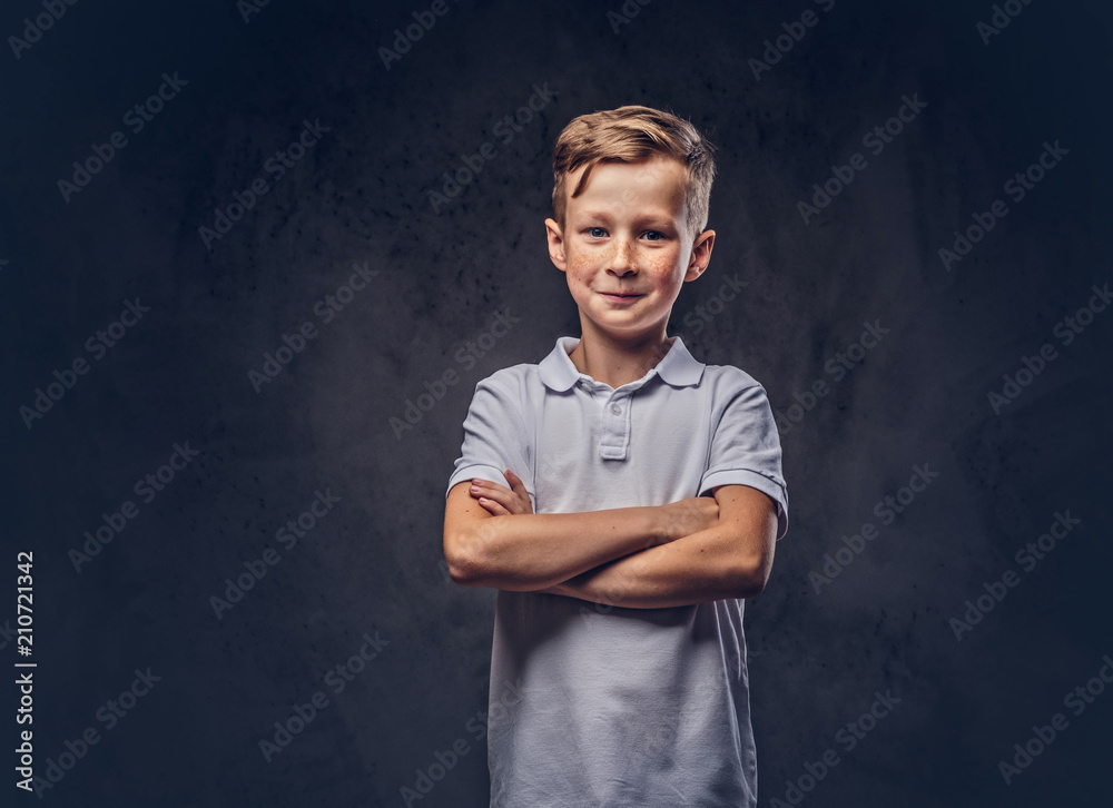 Cute little boy dressed in a white t-shirt standing with crossed arms in a studio. Isolated on dark textured background.