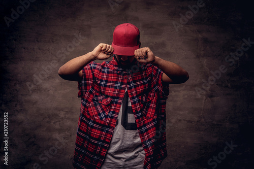 Portrait of an African-American dancer guy dressed in a red fleece shirt and cap at the studio. Isolated on dark textured background. © Fxquadro