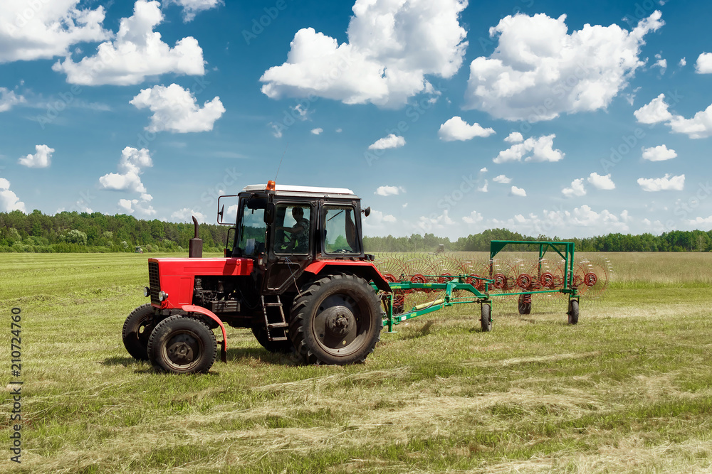 Agricultural machinery, a tractor collecting grass in a field against a blue sky. Hay harvesting, grass harvesting. Season harvesting, grass, agricultural land. Agricultural machinery, a tractor collecting grass in a field against a blue sky. Hay harvesting, grass harvesting. Season harvesting, grass, agricultural land.