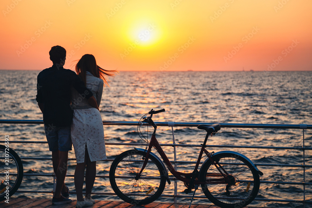 Couple of young hipster lovers hugging at the beach and watching the sunrise at wooden deck summer time