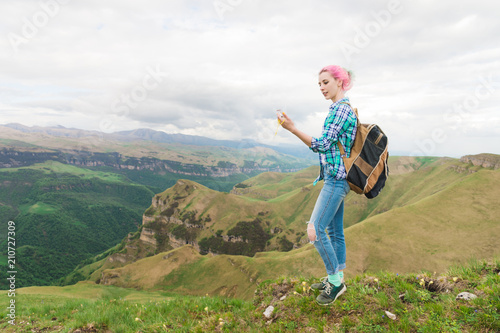 A hipster girl traveled by a blogger in a plaid shirt and with multi-colored hair using a compass standing on a plateau background in the mountains