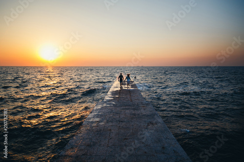 Young couple running at the pier towards the sunrise sun at the beach summer time