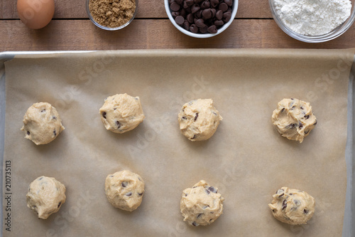 Flat lay of chocolate chip cookie dough balls on a baking sheet, with line of ingredient bowls