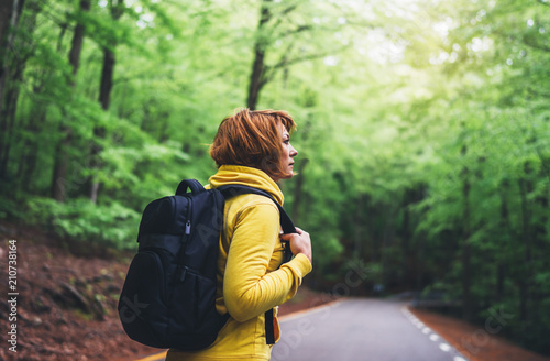 tourist traveler with backpack into road at summer green forest, girl hiker in yellow hoody looking and enjoying the breath of fresh clean air in trip, relax holiday concept, blurred background