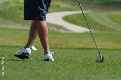 Young man leaning on his golf club on the putting green wearing blue shorts and white golf shoes. Playing golf in the summertime on a golf course.