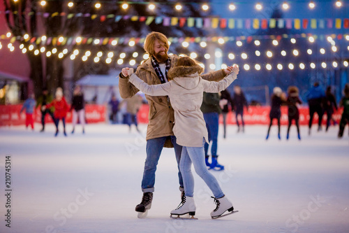 Theme ice skating rink and loving couple. meeting young, stylish people ride by hand in crowd on city skating rink lit by light bulbs and lights. Ice skating in winter for Christmas on ice arena