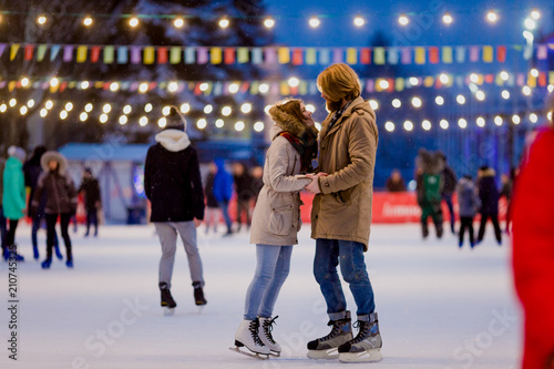 Ice skating rink and lovers together. A pair of young people in an embrace on a city skating rink lit by light bulbs and bright lights. Winter date for Christmas on the ice arena