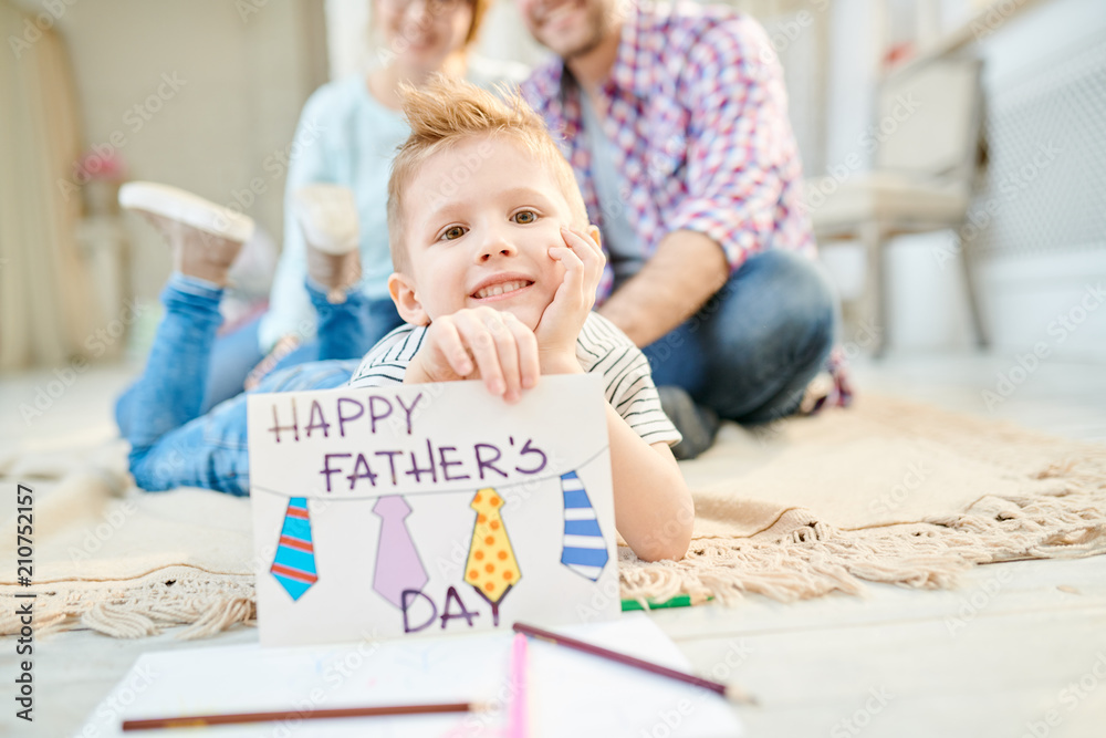 Warm toned portrait of cute little boy lying on floor in living room posing with handmade Fathers day card  and smiling looking at camera