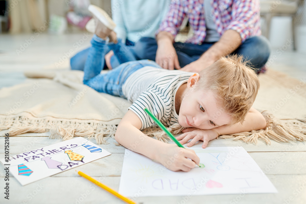 Warm toned portrait of cute little boy lying on floor in living room drawing picture for Fathers day for dad and smiling, copy space Warm toned portrait of cute little boy lying on floor in living room drawing picture for Fathers day for dad and smiling, copy space
