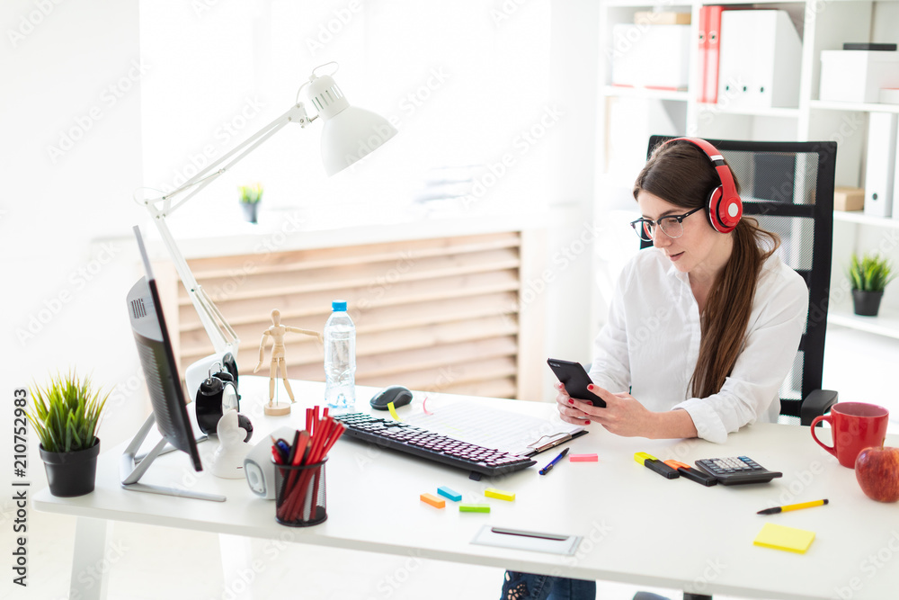 A young girl sits in headphones at a table in the office and holds a phone in her hand. A young girl sits in headphones at a table in the office and holds a phone in her hand.