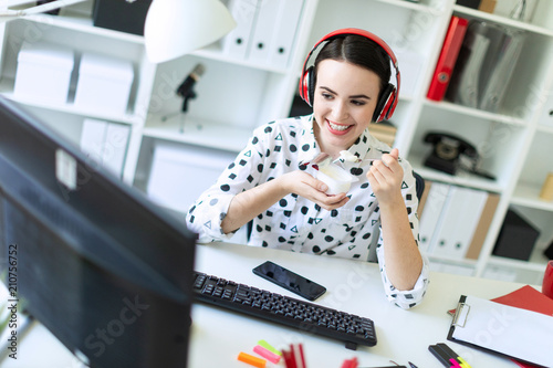 Beautiful young girl sitting in headphones at desk in office, eating yogurt and looking at monitor. photo