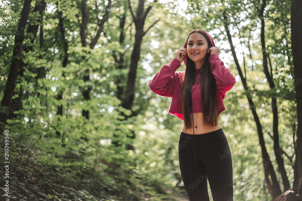 Beautiful sports woman put oh headphones before training outdoors. Portrait of a young athletic woman in pink hoodie put on headphones in the forest. Sports smiling female listening to music Beautiful sports woman put oh headphones before training outdoors. Portrait of a young athletic woman in pink hoodie put on headphones in the forest. Sports smiling female listening to music