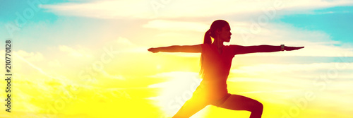 Yoga woman meditating zen in warrior pose relaxing outside by beach at sunrise or sunset. Female yoga instructor working out training in sun flare.