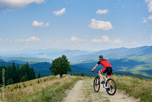 Back view of sportsman cyclist in sportswear and helmet riding cross country bicycle on mountain road. Carpathian mountains view and blue sky on background. Active lifestyle and outdoor sport concept