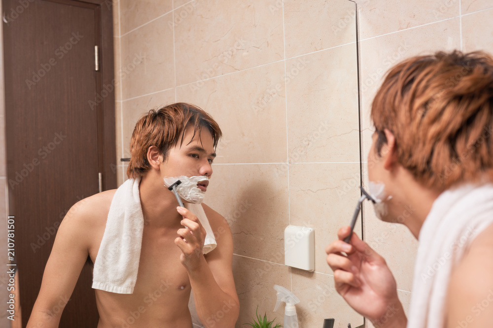 Handsome young asian man is shaving his face and looking at the mirror.
