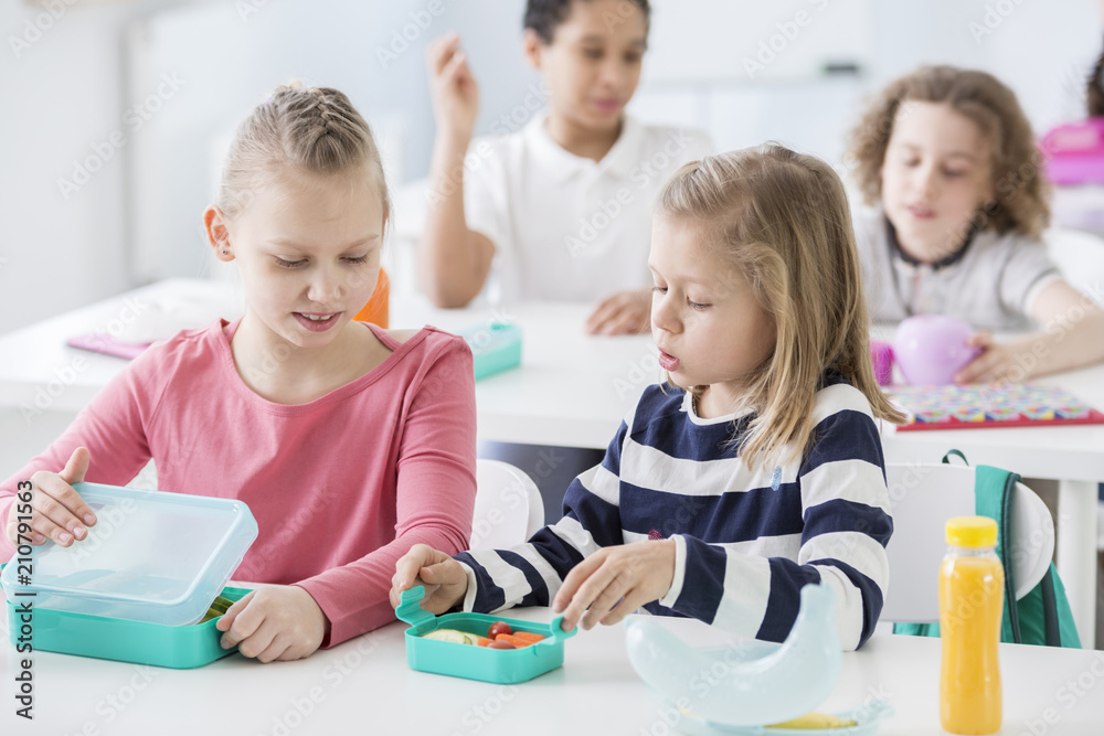 Snack time in a kindergarten class. Children opening their mint color lunch boxes with healthy vegetables. A bottle of orange juice on the desk. Snack time in a kindergarten class. Children opening their mint color lunch boxes with healthy vegetables. A bottle of orange juice on the desk.