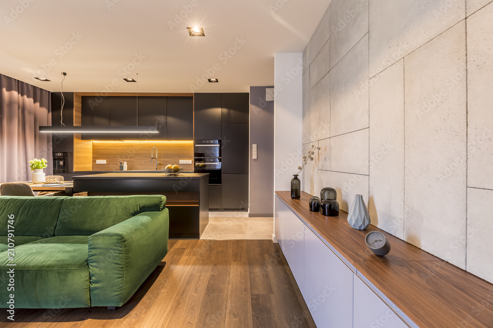 Wooden cupboard with vases and clock in modern elegant living room interior with green velvet sofa and dark kitchen corner Wooden cupboard with vases and clock in modern elegant living room interior with green velvet sofa and dark kitchen corner