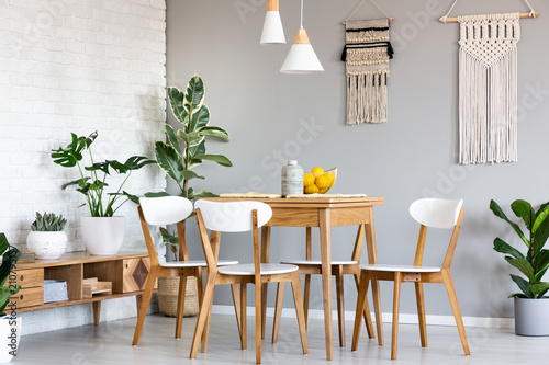 Macrame hanging on gray wall above wooden table and chairs in bright dining room interior with lots of plants. Real photo