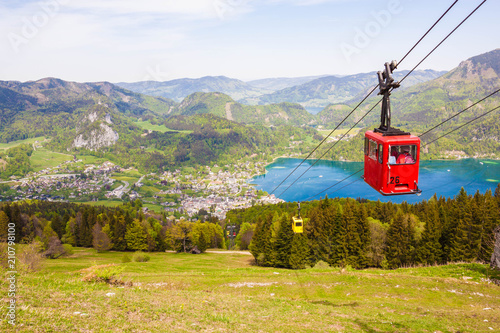 Yellow and red gondolas of Zwoelferhorn Seilbahn (cable way) travel up and down alpine peak with a view of village St.Gilgen, Wolfgangsee lake, surrounding hills and mountains. Salzburg land, Austria photo