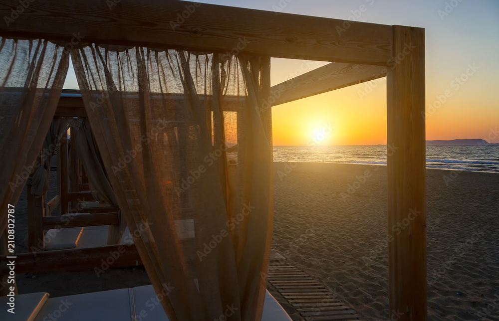 Silhouette wooden with blinds gazebo on an empty sandy beach on a sunset background.