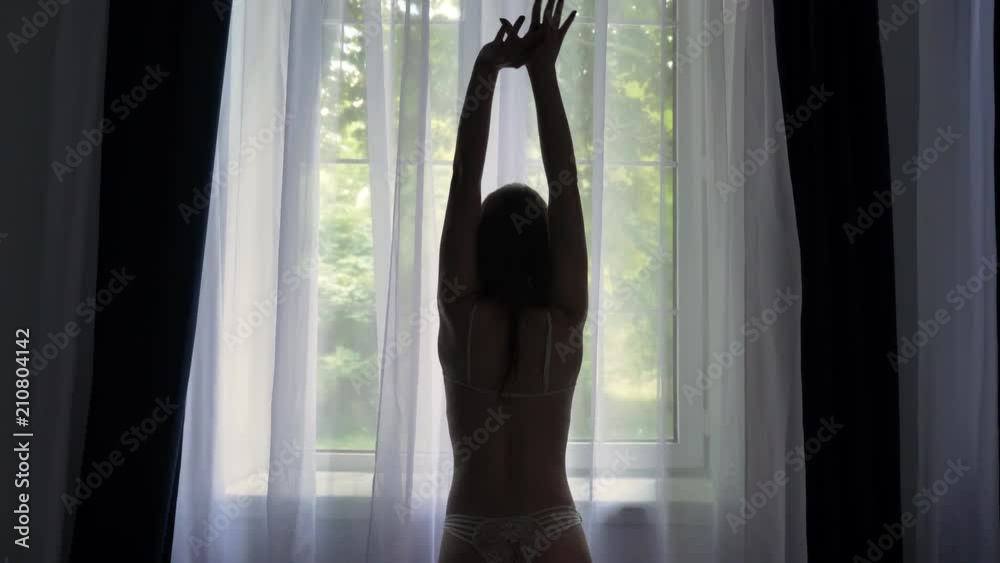 Silhouette of young sexy girl in lingerie is standing near window and raising hands, watching at camera, tulle on background, flirt concept Silhouette of young sexy girl in lingerie is standing near window and raising hands, watching at camera, tulle on background, flirt concept