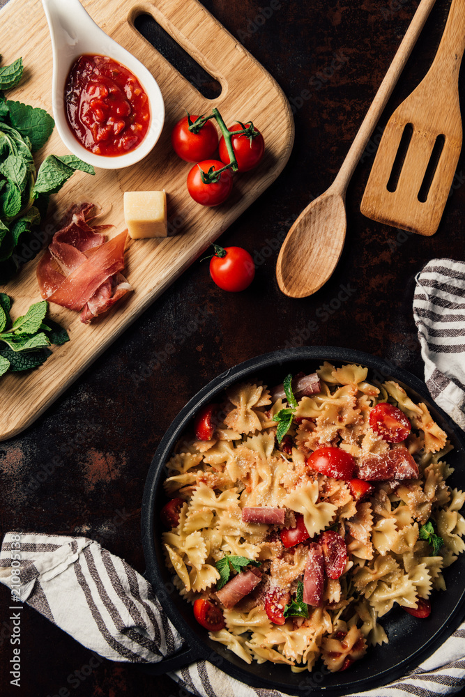 elevated view of pasta with jamon, pine nuts, sauce, cherry tomatoes, mint leaves covered by grated parmesan in pan surrounded by kitchen towel and cutting board with ingredients on table elevated view of pasta with jamon, pine nuts, sauce, cherry tomatoes, mint leaves covered by grated parmesan in pan surrounded by kitchen towel and cutting board with ingredients on table