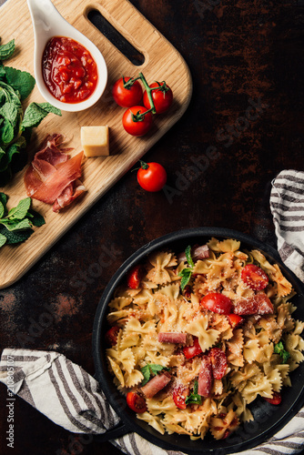 top view of pasta with jamon, pine nuts, sauce, cherry tomatoes, mint leaves covered by grated parmesan in pan surrounded by kitchen towel and cutting board with ingredients on table