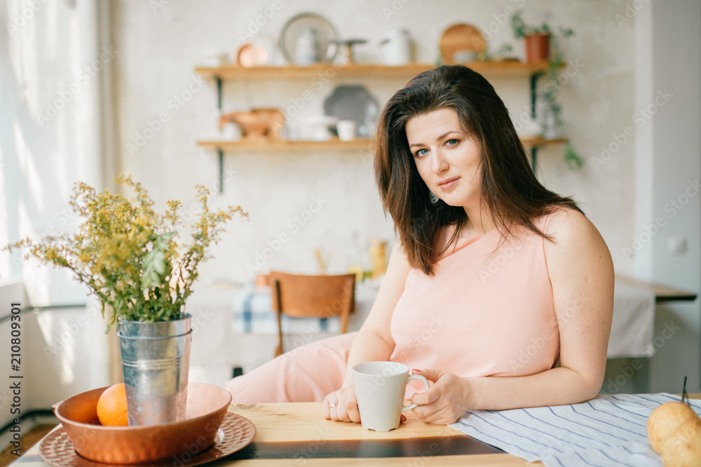 Young beautiful housewife in dress having breakfast on kitchen. Lovely woman posing and dreaming in country french style decorated interior zone. Rustic dining room with fresh food and tableware. Young beautiful housewife in dress having breakfast on kitchen. Lovely woman posing and dreaming in country french style decorated interior zone. Rustic dining room with fresh food and tableware.